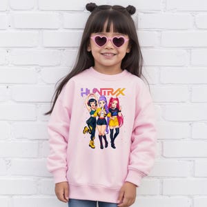 May include: A light pink sweatshirt featuring a colorful graphic of three anime-style figures with the text "HUNTRAX" in purple and yellow. The figures are in various poses, wearing different outfits. The sweatshirt is worn by a young girl wearing heart-shaped sunglasses.