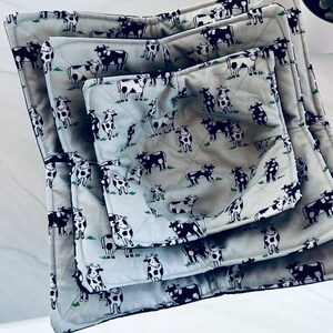 May include: Set of three quilted bowl cozies in varying sizes. The light gray fabric features a repeating pattern of black and white cows on a green grassy background. The cozies are designed to insulate bowls and protect hands from heat.