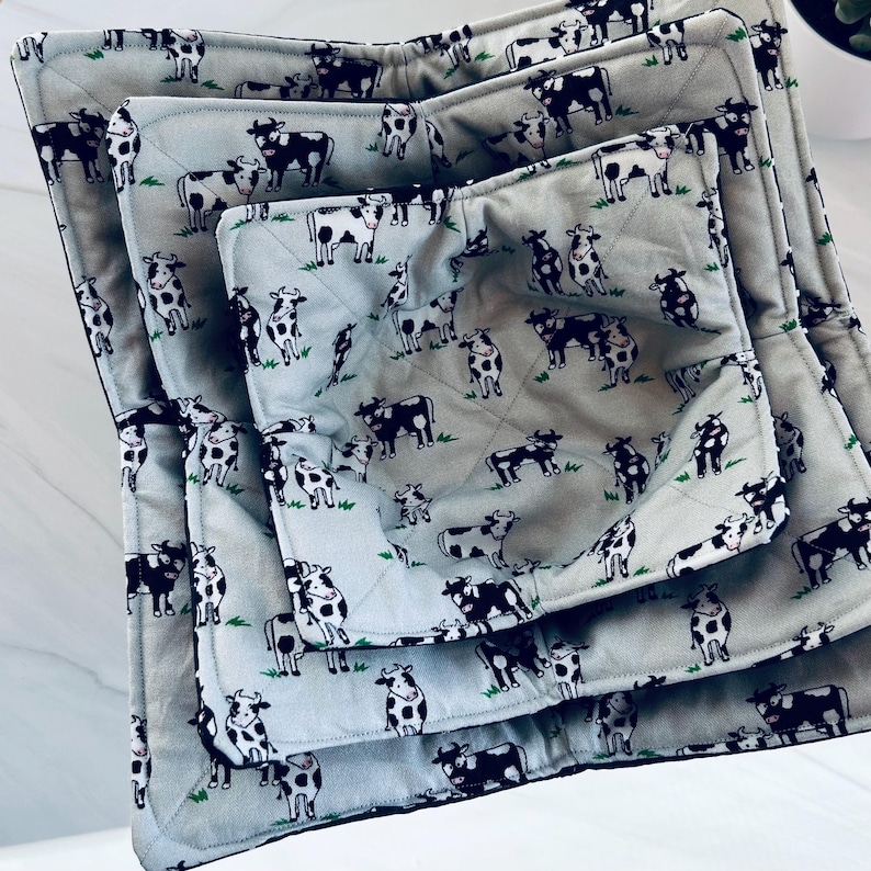 May include: Set of three quilted bowl cozies in varying sizes. The light gray fabric features a repeating pattern of black and white cows on a green grassy background. The cozies are designed to insulate bowls and protect hands from heat.