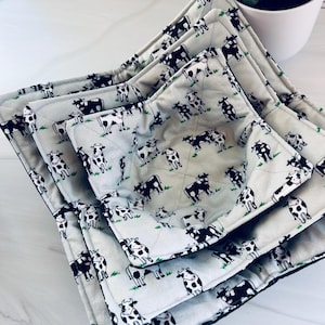 May include: A set of four gray bowl cozies with a repeating pattern of black and white cows grazing on green grass. The cozies are quilted and have a black trim.