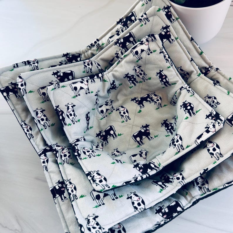May include: A set of four gray bowl cozies with a repeating pattern of black and white cows grazing on green grass. The cozies are quilted and have a black trim.