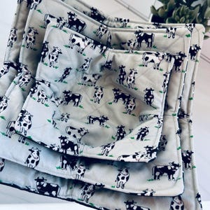May include: A set of three quilted pot holders and a matching oven mitt. The light gray fabric is patterned with black and white cows and green grass. The items are arranged in a nested fashion, showcasing the different sizes.