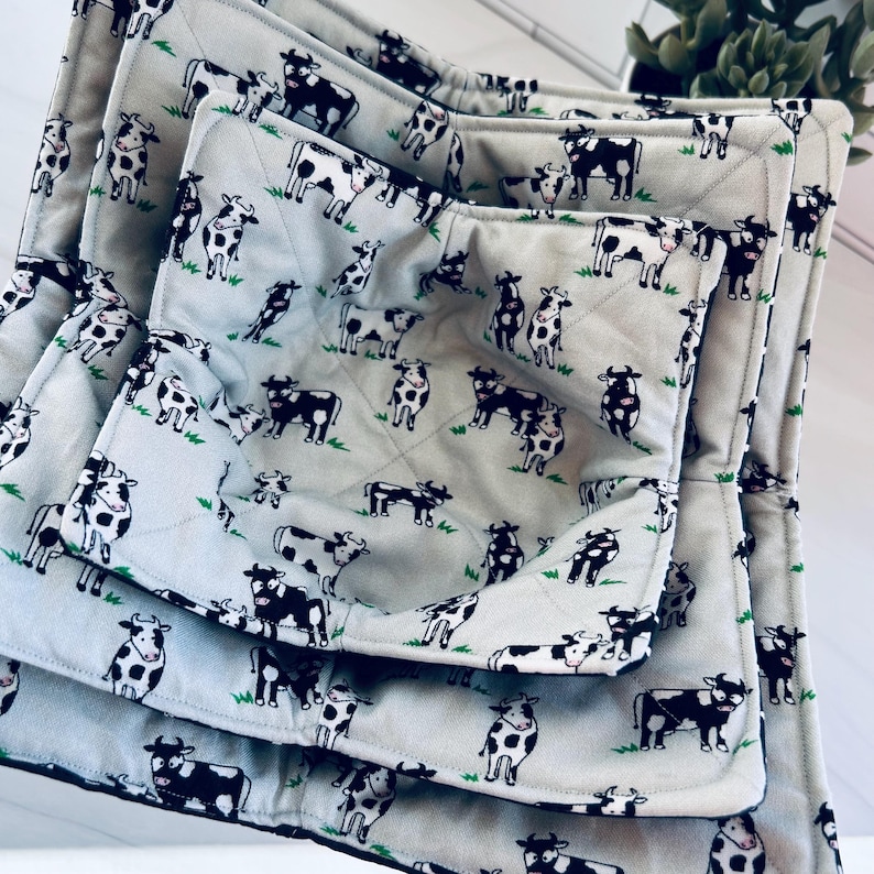 May include: A set of three quilted pot holders and a matching oven mitt. The light gray fabric is patterned with black and white cows and green grass. The items are arranged in a nested fashion, showcasing the different sizes.