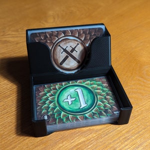 May include: A black card holder with two decks of cards. The top deck has a brown card with a crossed swords symbol. The bottom deck has a green card with a +1 symbol.