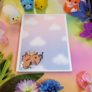 May include: A white notepad with a cartoon illustration of a capybara with a group of cats and a bird on its back. The background is a light blue and pink gradient with white clouds.