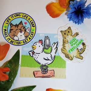 May include: Three stickers featuring cartoon cats. One sticker says "Life is long - Life is crap - Make it better - Get a cat". Another sticker shows a cat riding a chicken on a spring. The third sticker shows a cat with the text "100% That Freeloader".