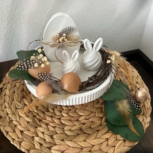Könnte beinhalten: Eine österliche Dekoration mit einem weißen, eiförmigen Behälter mit Federn und Bindfaden, zwei weißen Hasenfiguren und braunen Eierschalen. Die Anordnung befindet sich auf einem weißen Tablett mit einer gewebten Tischset-Basis, akzentuiert mit Federn und Grün.