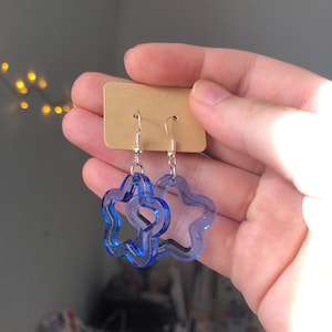 May include: A pair of blue, flower-shaped earrings. The earrings are made of a transparent material and have a silver hook. The earrings are displayed on a small, brown cardboard tag. The background is a blurred, neutral colour.