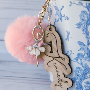 May include: A pink fluffy pom-pom keyring with a gold ballerina charm and a wooden tag with the name "Kennedy" engraved on it.
