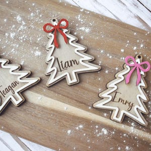 以下が含まれることがあります： 「Liam」、「Teagan」、および「Emry」の名前が刻まれた3つの木製のクリスマスツリーの装飾品。白い粉をまぶした木製の板の上に置かれ、各ツリーには赤またはピンクのリボンが付いています。