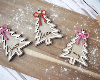 Peut inclure: Trois décorations de Noël en forme de sapin en bois, avec les noms "Liam", "Teagan" et "Emry". Elles sont posées sur une planche de bois saupoudrée de poudre blanche, avec un nœud rouge ou rose.