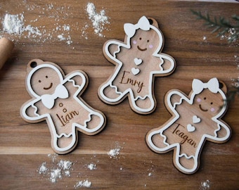 May include: Three wooden gingerbread person ornaments with white outlines. The ornaments are decorated with pink hearts and have the names "Liam", "Emry", and "Teagan" written on them.