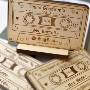 Könnte beinhalten: Ein helles Holzornament in Form einer Kassette auf einem Ständer, graviert mit "Third Grade mix Vol 3", "Ms. Bartoli" und einem Spotify-Code. Das Design enthält Herz- und Sternsymbole. Zwei weitere ähnliche Holzornamente sind darunter zu sehen.