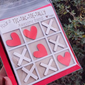 Puede incluir: Juego de tres en raya con corazones rojos y cruces blancas. El juego está en una funda de plástico transparente con el texto "Your a TIC-TAC-TOE-TALLY Awesome friend".