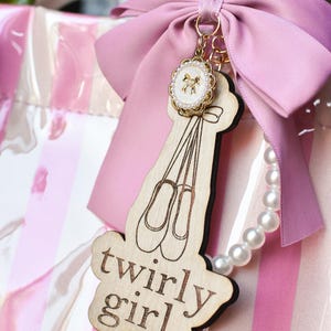 May include: A wooden keychain with a ballet shoe design and the words "twirly girl." The keychain is attached to a pink bow and a pearl necklace. A gold-colored charm with a bow design is also attached.