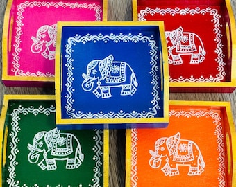 May include: Five wooden serving trays with painted elephant designs. The trays are in pink, blue, green, orange, and red. Each tray has a gold border and a white elephant design.