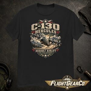 Puede incluir: Una camiseta negra con un gráfico de un avión C-130 Hercules sobrevolando montañas. El texto dice C-130 HERCULES, ASSAULT AIRLIFT y LOW. LOUD. LEGENDARY. El diseño incluye un emblema de estrella y detalles en colores rojo, blanco y azul.