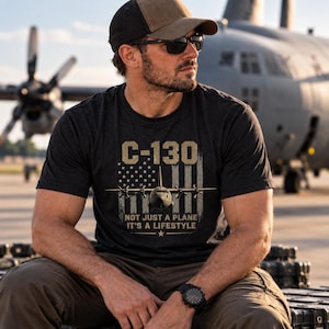 May include: A charcoal grey short-sleeve t-shirt featuring a graphic of a C-130 aircraft centred over a distressed American flag design. The text reads C-130 NOT JUST A PLANE IT'S A LIFESTYLE with a small star icon below the lettering.