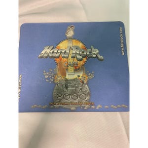 May include: A Hard Rock Cafe coaster with a blue background. The coaster features the Hard Rock Cafe logo with a guitar graphic and the text "2000 THE EVOLUTION OF ROCK". The website address www.hardrock.com is printed on the coaster.