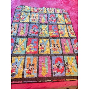 Pode incluir: Um edredom colorido com o alfabeto, cada letra em uma cor diferente. Cada quadrado de letra apresenta um personagem da Disney, incluindo Mickey Mouse, Minnie Mouse e Pluto. O edredom tem uma borda preta e está em uma superfície rosa.