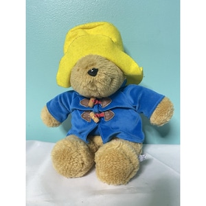 Puede incluir: Un peluche de Paddington Bear, con un sombrero amarillo y un abrigo azul con cierres de palanca de madera. El oso tiene un cuerpo marrón y una nariz negra. El juguete está sobre una superficie blanca contra un fondo azul claro.