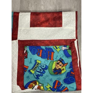 May include: A handmade quilt featuring a colorful design. The quilt has a red and white patchwork pattern with a central panel showcasing a cartoon character and the word "HERO". The quilt is likely for children.