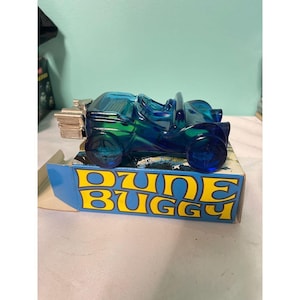 Puede incluir: Un coche de juguete Dune Buggy azul de plástico transparente. El coche está sobre una caja con las palabras "Dune Buggy" en amarillo y azul. El coche tiene un diseño detallado con ruedas visibles y una sección trasera.