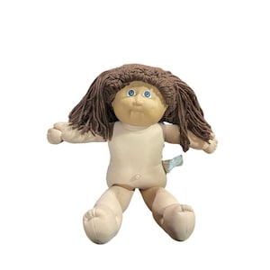 May include: A vintage Cabbage Patch Kid doll with a tan skin tone, blue eyes, and brown yarn hair. The doll is wearing a tan body suit and has its arms and legs outstretched. A small tag is attached to the doll's body.