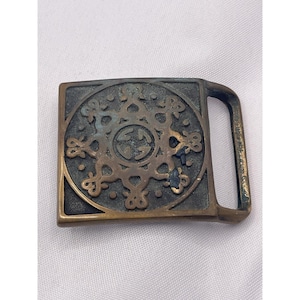 May include: Vintage brass belt buckle with a square shape and a decorative circular design in the center. The design includes a logo and ornate floral-like patterns. The buckle has a rectangular bar for the belt.