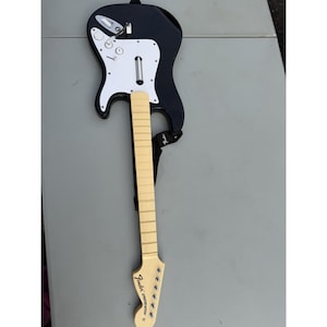 Może przedstawiać: Czarno-biały kontroler do gier wideo Fender Stratocaster. Kontroler posiada błyszczący czarny korpus, białą maskownicę, jasnobrązową szyjkę z oznaczeniami progów oraz czarny pasek z nadrukowanym logo Fender.