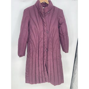 May include: A long, quilted coat in a deep plum color. The coat features a high collar, button closures, and vertical stitching details. The coat is hanging on a wooden hanger.