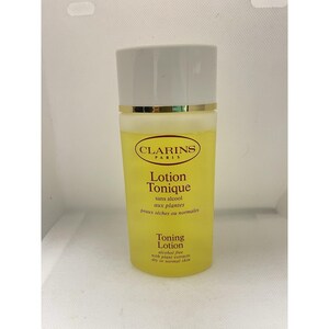Puede incluir: Una botella amarilla de Clarins Lotion Tonique con tapa blanca y borde dorado. La botella tiene texto en francés e inglés, incluyendo "Lotion Tonique" y "Toning Lotion". El producto es sin alcohol y para pieles secas o normales.