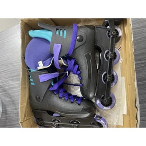 Puede incluir: Patines en línea negros con cordones y detalles morados, y ruedas moradas. Los patines están en una caja de cartón. La bota tiene un exterior negro con acolchado turquesa y morado.