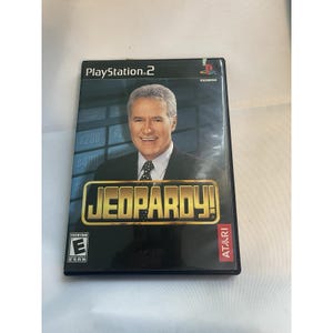 May include: A PlayStation 2 video game case for Jeopardy! features a portrait of the show's host, Alex Trebek. The game's title is in gold lettering. The case is black with the ESRB rating and Atari logo.