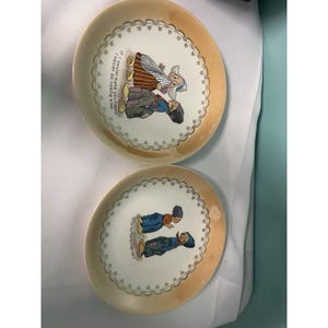 Peut inclure: Deux assiettes décoratives vintage avec un centre crème et un bord pêche. Chaque assiette présente une illustration de personnages en costumes traditionnels hollandais. Du texte est visible sur chaque assiette.