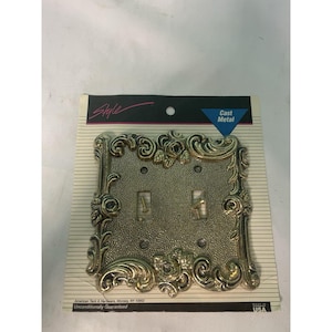 Puede incluir: Cubierta de interruptor de luz doble dorada ornamentada con un diseño floral y de volutas detallado. Las palabras "Style" y "Cast Metal" son visibles. Hecho en los EE. UU.