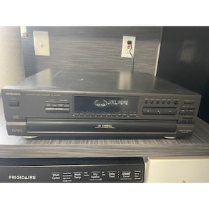Könnte beinhalten: Ein grauer Technics CD-Wechsler SL-PD707. Die Frontplatte verfügt über eine Digitalanzeige mit Titelinformationen und Bedientasten. Das Gerät hat eine Kapazität für 5 CDs und das Wort "MASH" ist auf der Vorderseite aufgedruckt.