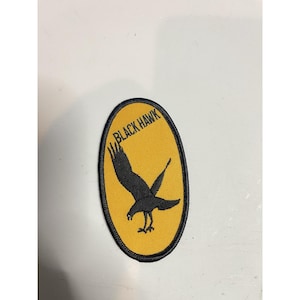 Puede incluir: Parche bordado con un halcón negro sobre un óvalo amarillo. El texto "BLACK HAWK" está arqueado sobre el ave. El parche tiene un borde negro. Ideal para chaquetas, bolsos u otros artículos de tela.