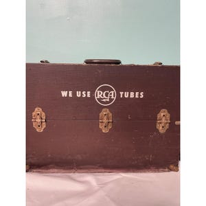 May include: A dark brown wooden box featuring the text "WE USE RCA TUBES" in white. The box has three metal clasps and a handle. The box appears to be vintage and shows signs of wear.