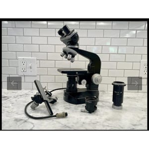 May include: A black Nikon microscope with silver-colored lenses and a stage. Additional components include a light source, an eyepiece, and a lens. The microscope is set against a white tile background.