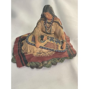 Puede incluir: Una figura detallada que representa a una mujer nativa americana sosteniendo a un bebé. La mujer viste un vestido color canela, collares de cuentas y un tocado negro. El bebé está en una cuna. La base es una mezcla de rojo, verde y marrón.