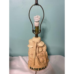 May include: Cream-colored baseball-themed lamp with a baseball bat, glove, ball, and cap design. The base reads "BASEBALL" in black lettering. The lamp has a brass-colored neck and a white spiral lightbulb.