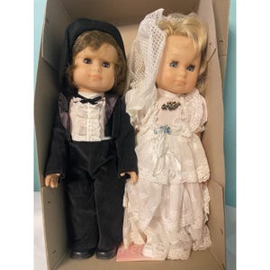 May include: Two vintage dolls in a cardboard box. One doll is dressed as a groom in a black tuxedo with a black hat. The other doll is dressed as a bride in a white lace wedding dress and veil. Both dolls have blonde hair.