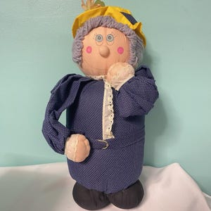 Puede incluir: Una muñeca de tela vintage con una cara sonriente, vestida con un vestido azul con lunares, un sombrero amarillo y cabello gris. La muñeca tiene mejillas rosadas y se lleva una mano a la cara. La muñeca está sobre una superficie blanca.