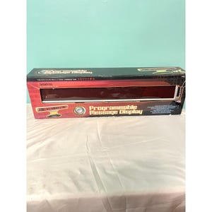 Puede incluir: Caja rectangular para una pantalla de mensajes programable. La caja es negra y roja con las palabras "Programmable Message Display" en blanco. La caja tiene una ventana transparente para ver la pantalla.