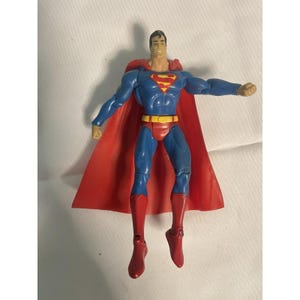 May include: A Superman action figure in a flying pose. The figure is wearing a blue suit with a red "S" emblem on the chest, red briefs, and red boots. A red cape flows behind the figure. The figure has a yellow belt and black hair.