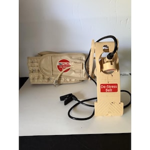 Puede incluir: Un sistema de cinturón De-Stress Belt beige que incluye una faja acolchada ancha con un logotipo circular y una unidad de bomba mecánica vertical conectada. La bomba tiene una manguera negra, un clip negro y una etiqueta roja con el texto.