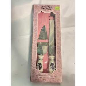May include: A porcelain wedding cake knife and server set by Enesco. The set includes a cake knife and server with silver blades and white handles with a wedding scene design. The box is pink with a rose pattern.