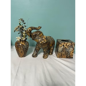 May include: A decorative set featuring a ceramic elephant figurine, a vase with faux flowers, and a tissue box cover. The items have a brown and tan zebra stripe pattern. The vase is approximately 8 inches tall.