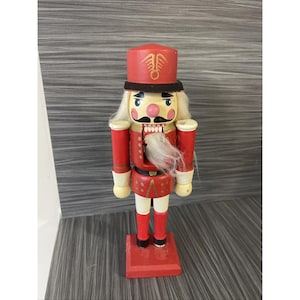 Puede incluir: Figura de cascanueces de madera con uniforme de soldado. La figura tiene sombrero y chaqueta rojos, pelo y barba blancos y bigote negro. El cascanueces se apoya sobre una base roja y es una decoración navideña clásica.